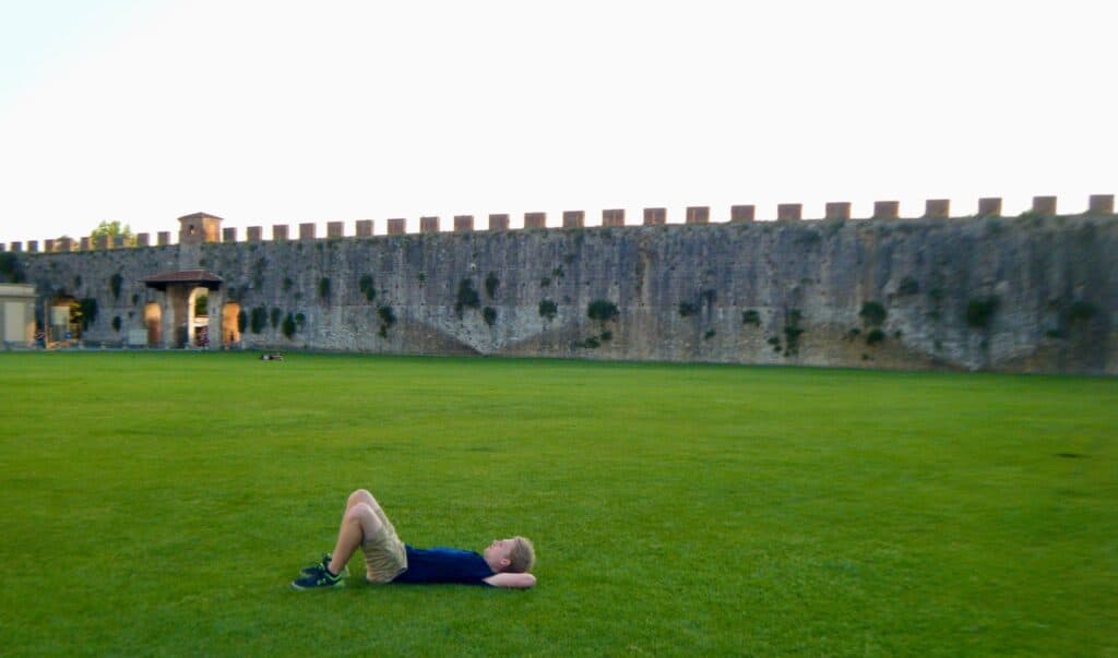 A man lies in manicured grass with his hands behind his head. An old strong wall surrounds the courtyard.