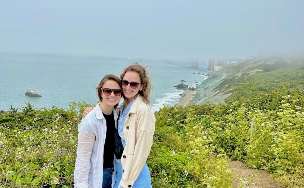 Two women stand in front of the Golden Gate Bridge which is largely obscured by clouds.