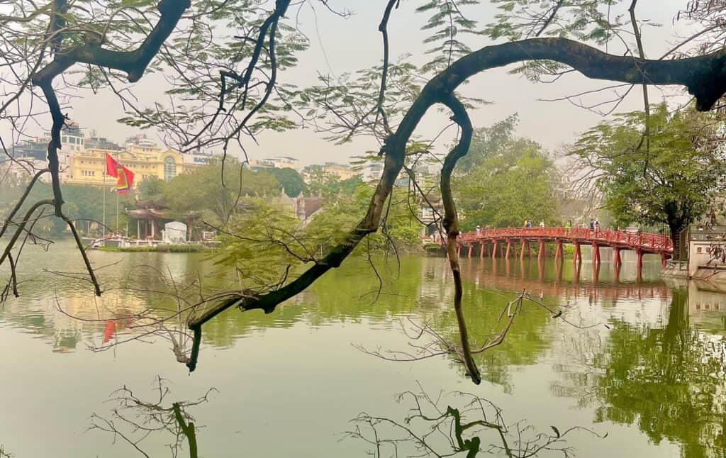 A scenic view of Hoan Kiem Lake in Hanoi, Vietnam, with a red wooden bridge leading to a small island. The lake is framed by tree branches, and in the background, there's a Vietnamese flag and city buildings visible.