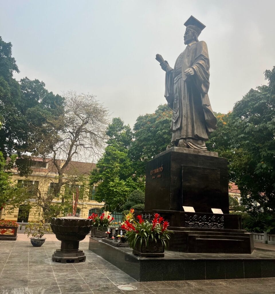 A large statue of Lý Thái Tổ, a historical figure dressed in traditional robes and holding a scroll, stands prominently in an open courtyard surrounded by trees. The statue is set on a tall pedestal with red flowers and offerings placed below it.