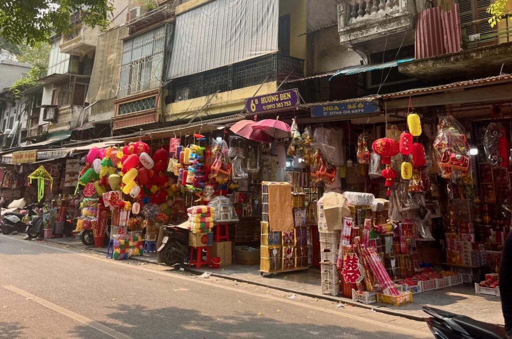 A colorful storefront in Vietnam displays an assortment of festive decorations including vibrant lanterns, banners, and paper goods. The street scene is lively, with various goods for sale spilling onto the sidewalk under awnings.