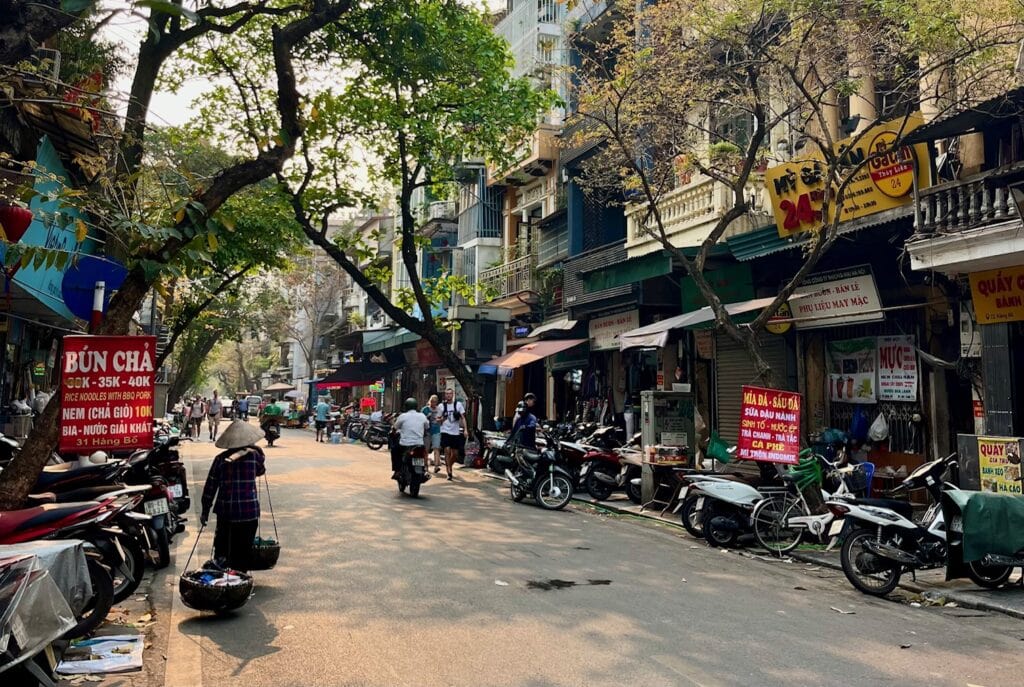 A bustling street in Hanoi during daylight, lined with trees and a variety of small shops and restaurants. A woman wearing a conical hat carries baskets using a bamboo pole, while motorcycles are parked along the roadside. A red sign advertises "Bún Chả" and other Vietnamese dishes.