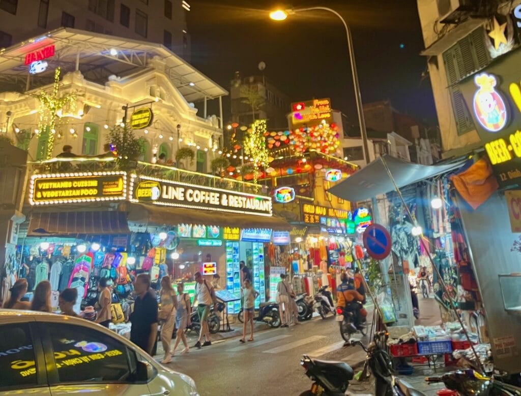 A lively street in Hanoi at night, brightly lit with neon signs for cafes, restaurants, and shops. Crowds of people walk along the street, with motorcycles and cars in the foreground. The scene is vibrant with colorful lights and bustling activity.