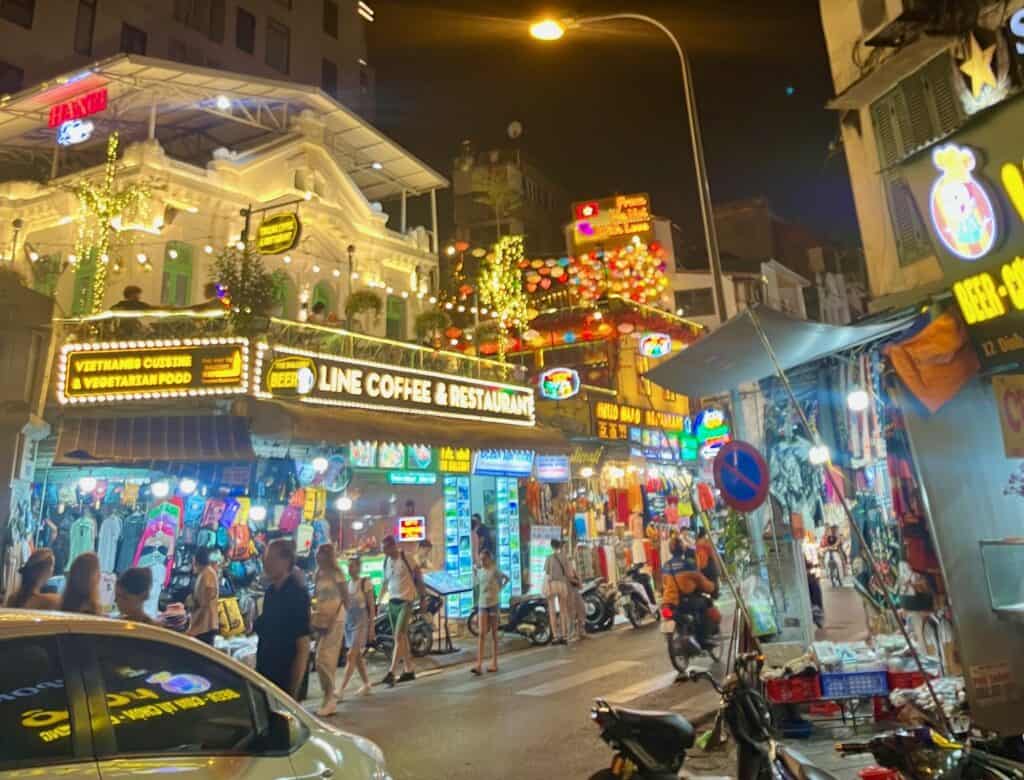 A vibrant night scene on a busy street in Vietnam, with bright neon signs advertising a coffee and restaurant, and a variety of shops selling clothes and souvenirs. The street is crowded with people, some walking and others on motorbikes, while festive lights and decorations adorn the buildings.