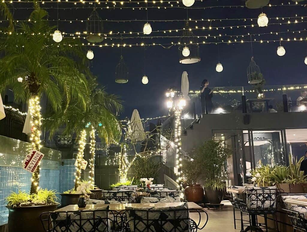 A rooftop restaurant at night, adorned with twinkling string lights and decorative hanging lamps. The area features palm trees wrapped in lights, tables and chairs arranged neatly, and a person standing on a higher platform, adding to the enchanting ambiance.