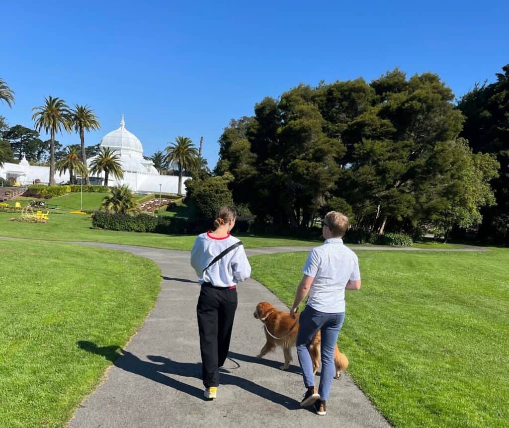 A couple walking their golden retriever on a sunny day in front of the white-domed Conservatory of Flowers in San Francisco's Golden Gate Park. The clear blue sky, palm trees, and well-manicured green lawn create a peaceful, leisurely scene.