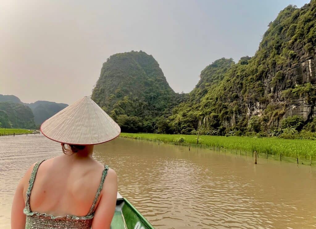 Is Vietnam safe for Solo Female travelers? A solo woman looks out at a muddy river surrounded by rice fields. She is wearing a traditional Vietnamese hat.