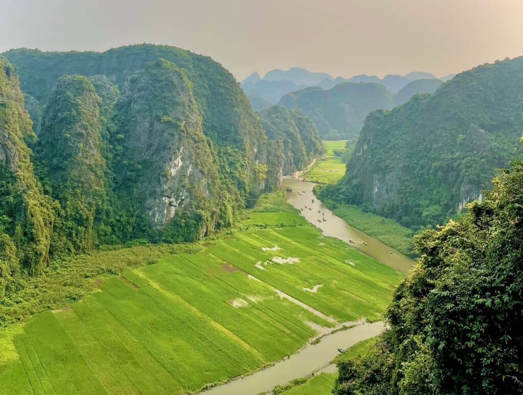 An aerial view of limestone mountains tower over green rice fields with a brown colored river running through the fields.