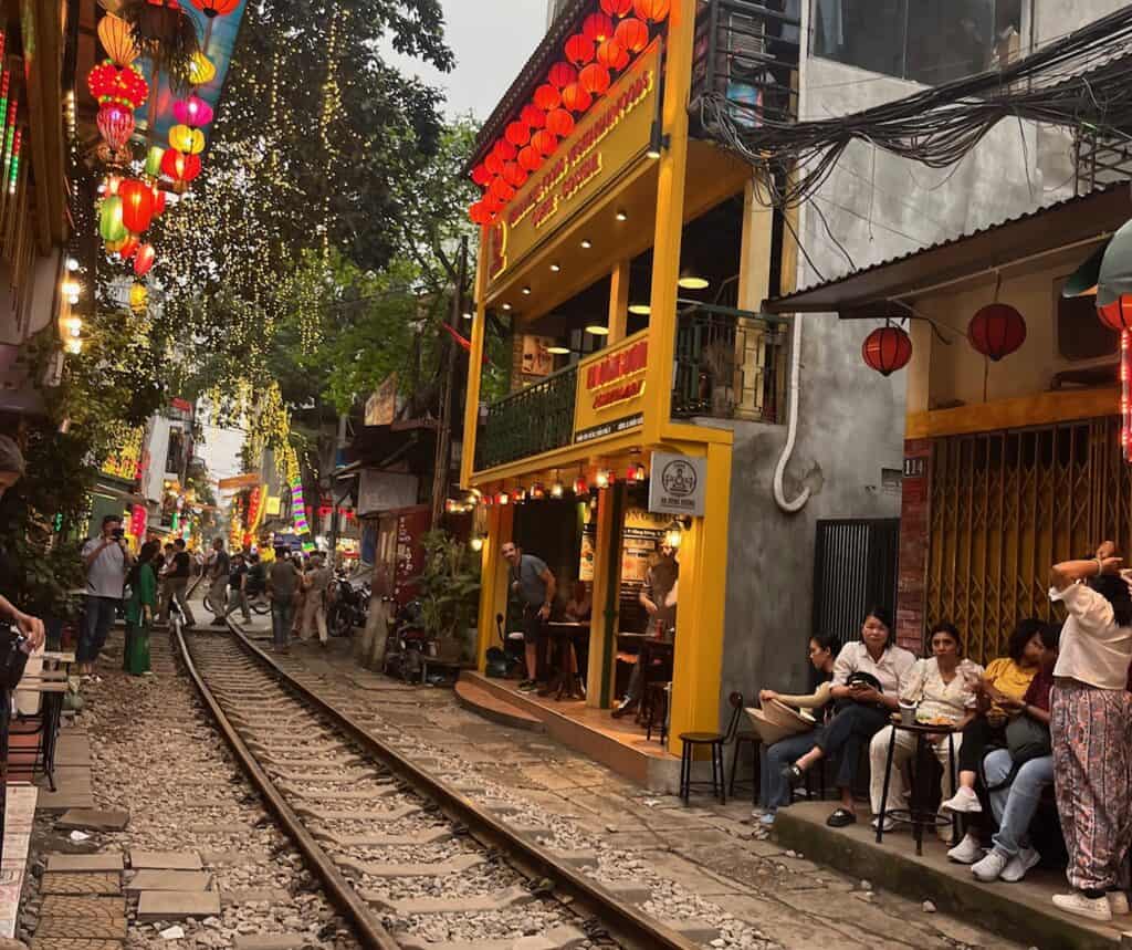 A narrow street in Hanoi, Vietnam, with train tracks running down the middle, surrounded by brightly lit lanterns and vibrant shops. People are sitting at outdoor cafes and walking along the tracks, creating a lively atmosphere."