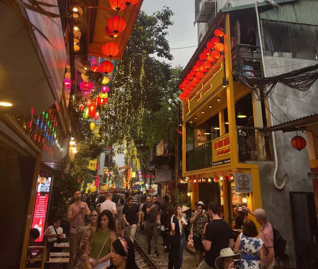 A crowded alleyway in Hanoi, Vietnam, decorated with hanging lanterns and twinkling lights. People walk along the railway track in the center of the alley, flanked by cafes and shops with vibrant signs, creating a lively and festive atmosphere.