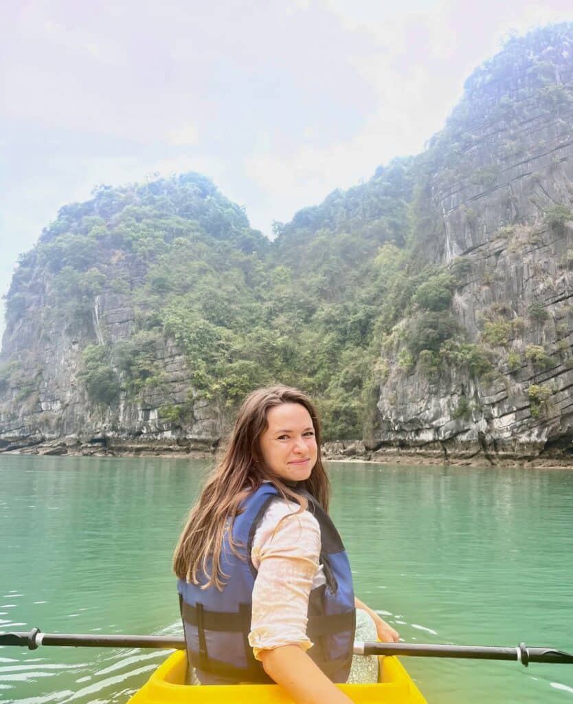 A smiling woman wearing a life jacket sits on a yellow kayak in turquoise waters with limestone cliffs surrounding the water.