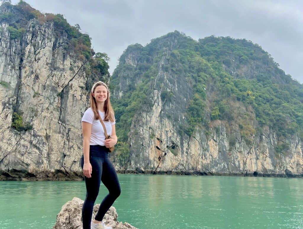 A solo woman stands on a rock in Halong Bay Vietnam with turquoise water and limestone cliffs behind her.