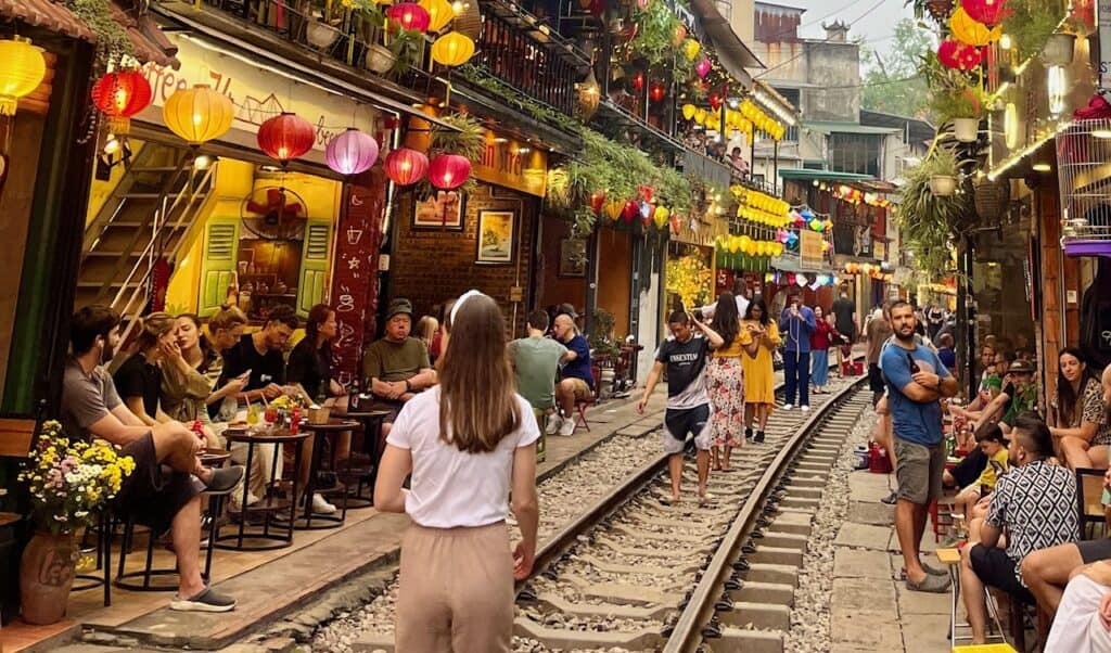 A lively cafe scene along a railway track in Hanoi, Vietnam, where people are seated at small tables, enjoying drinks and conversation. The narrow street is lined with colorful lanterns and bustling with visitors walking along the tracks, capturing the atmosphere of this unique location.