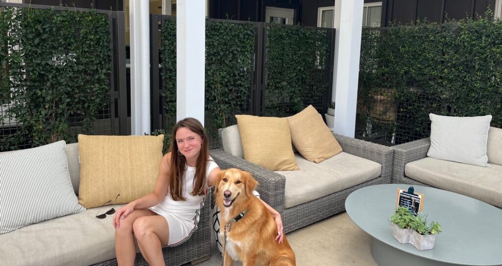 A woman in a white dress with her arm around the shoulders of a golden retriever. The woman is seated on a patio sofa.