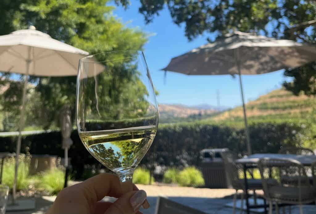 A hand holding a glass of white wine on a patio with tables and umbrellas as well as Napa vineyards in the background.