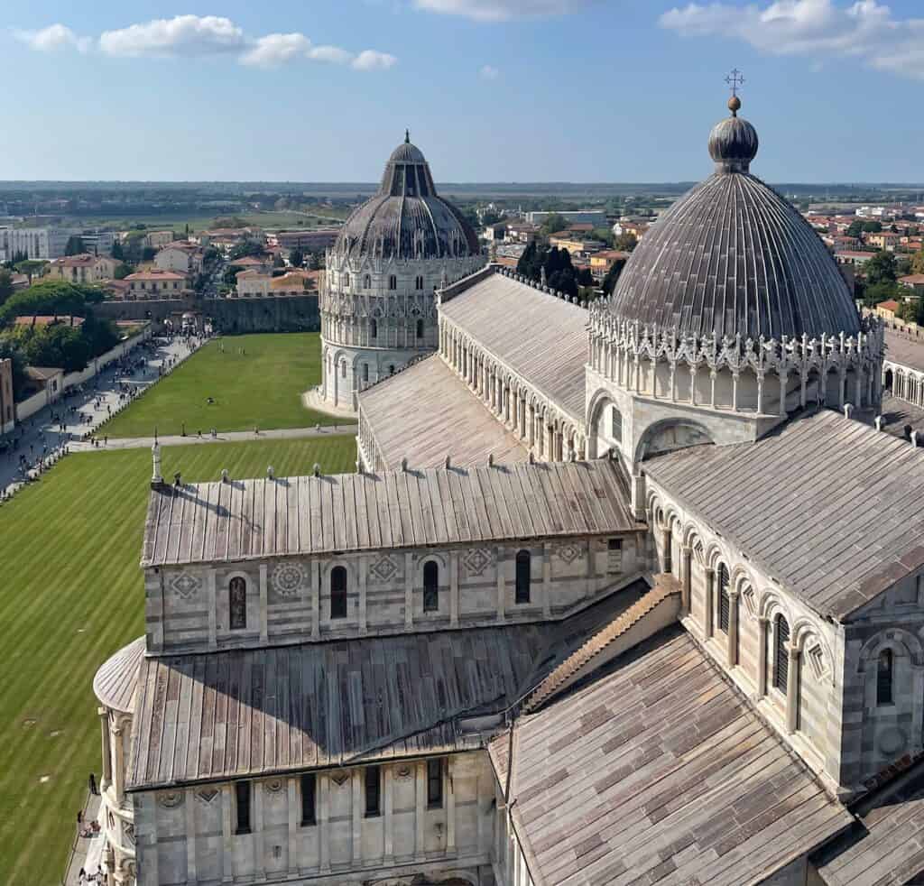 An aerial view of the Pisa Cathedral complex, showcasing the dome and rooftops of the Cathedral and Baptistery, with the surrounding landscape and distant city visible under a bright blue sky.