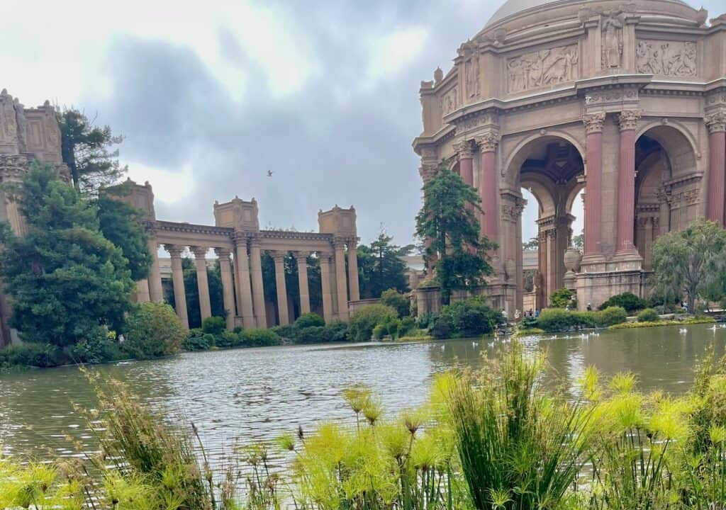 A large archway with ornate columns at the Palace of Fine Arts in San Francisco. A few people, including a man in an orange shirt holding a bike, are walking through the arch. The bright sky and surrounding greenery enhance the scene's grandeur. San Francisco 2 Day Itinerary should include the domed buildings of the Palace of Fine Arts.