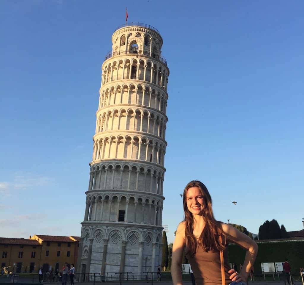 Woman in brown tank top stands in front of the Leaning Tower of Pisa at sunset.