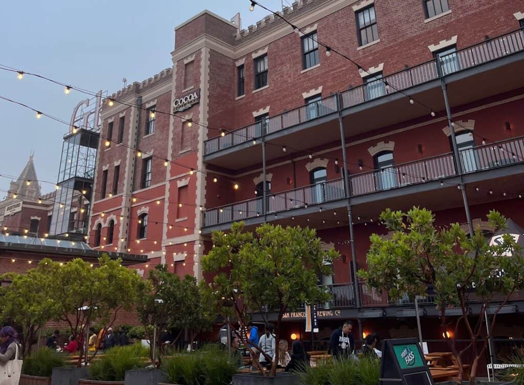 An outdoor evening scene at the San Francisco Brewing Co., featuring a red-brick building with string lights hanging overhead. People are seated at tables in the courtyard, surrounded by greenery. The building has a sign for "Cocoa" on the upper floor and a balcony running along its length.