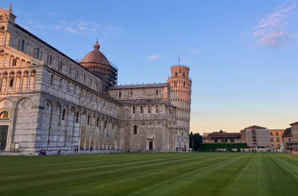 A side view of the Leaning Tower of Pisa alongside the Pisa Cathedral, with the sun setting, casting a golden hue on the buildings, and a well-manicured green lawn in the foreground.