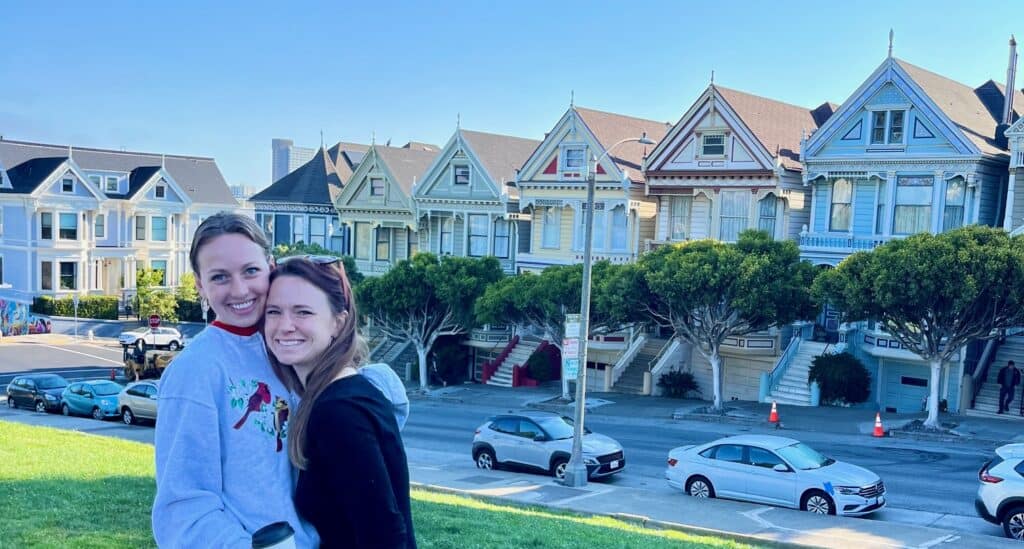 Two women smiling and hugging with the Painted Ladies Victorian houses behind them.