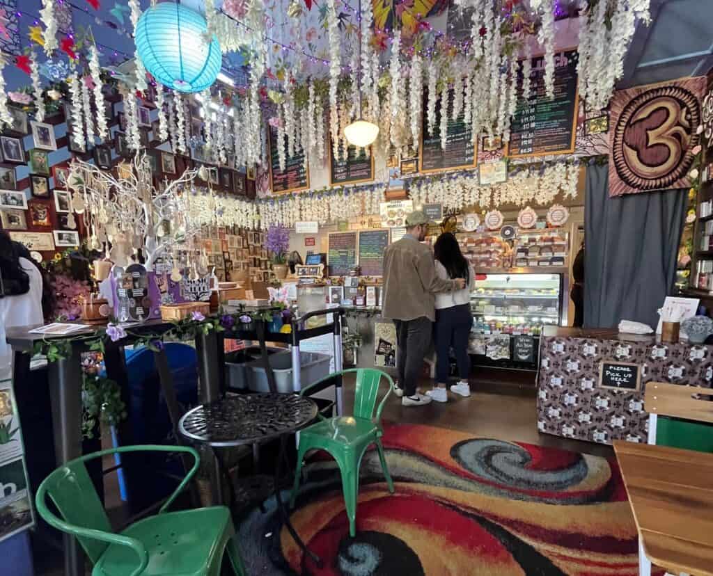  The interior of a cozy, eclectic cafe adorned with hanging flowers, picture frames, and colorful decorations. Two people stand at the counter looking at the menu, surrounded by mismatched furniture, green chairs, and a vibrant atmosphere.