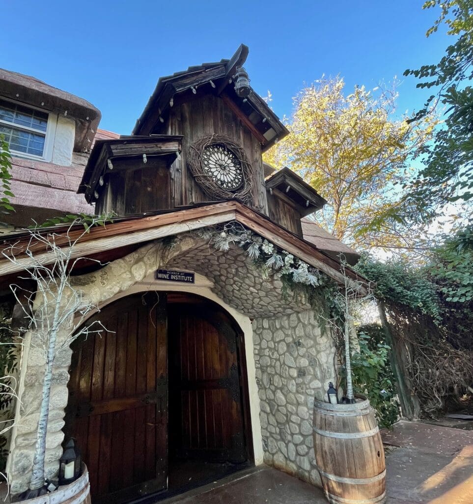 A close-up of a stone and wood entrance with a decorative circular window above the door. The door is arched and made of dark wood, with a sign above it that reads "Wine Institute." Two wooden barrels flank the entrance, and the building has a rustic, storybook-like appearance.