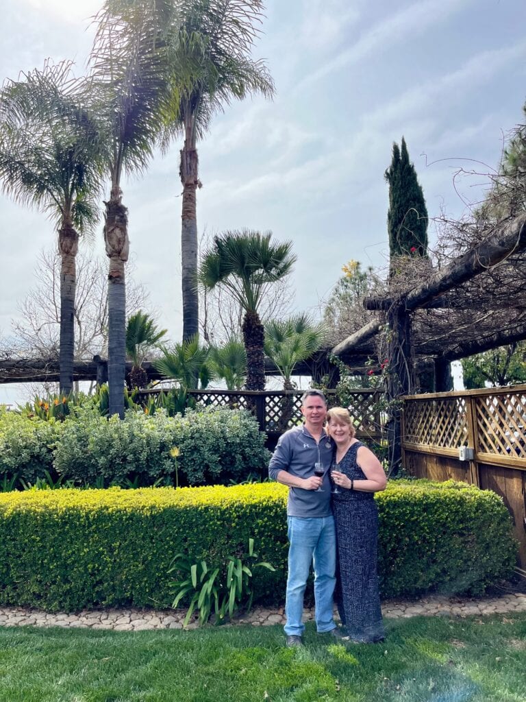 A couple standing in a lush, manicured garden with tall palm trees in the background. The couple, holding wine glasses, is smiling at the camera, surrounded by greenery and well-maintained hedges.