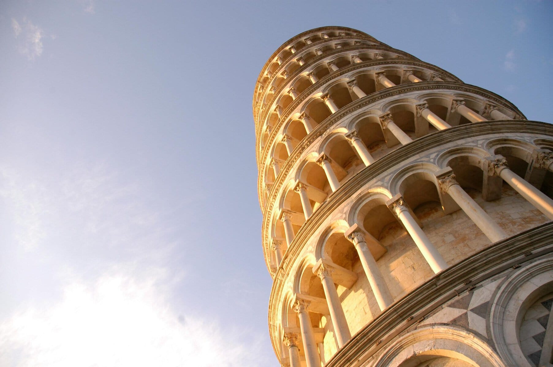 A low-angle view of the Leaning Tower of Pisa, focusing on its intricate arches and columns bathed in warm sunlight, with a clear blue sky in the background.