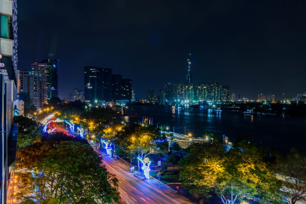 A panoramic night view of Ho Chi Minh City, showcasing brightly lit skyscrapers and buildings along the Saigon River. The city lights reflect on the water, and the streets are alive with streaks of light from traffic, highlighting the city's dynamic energy.