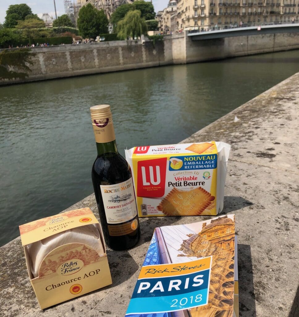 A small picnic setup by the Seine River in Paris includes a bottle of red wine, a box of Chavroux cheese, a pack of LU Petit Beurre biscuits, and a Rick Steves Paris guidebook. The backdrop includes historic buildings and part of the riverbank.