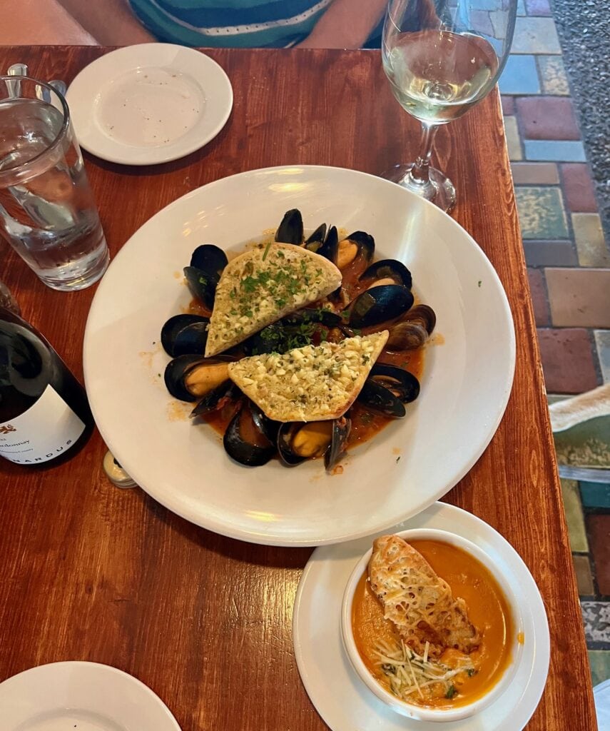 A seafood dish served on a wooden table, consisting of mussels in a savory sauce, topped with garlic bread. A bowl of creamy bisque and a glass of white wine accompany the main dish, alongside water and an empty plate.