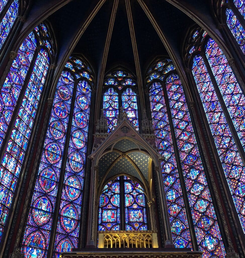 The interior of a Gothic-style cathedral with tall, intricate stained glass windows glowing in blue, red, and purple hues. The arched ceiling and ornate altar are visible below the vibrant windows.