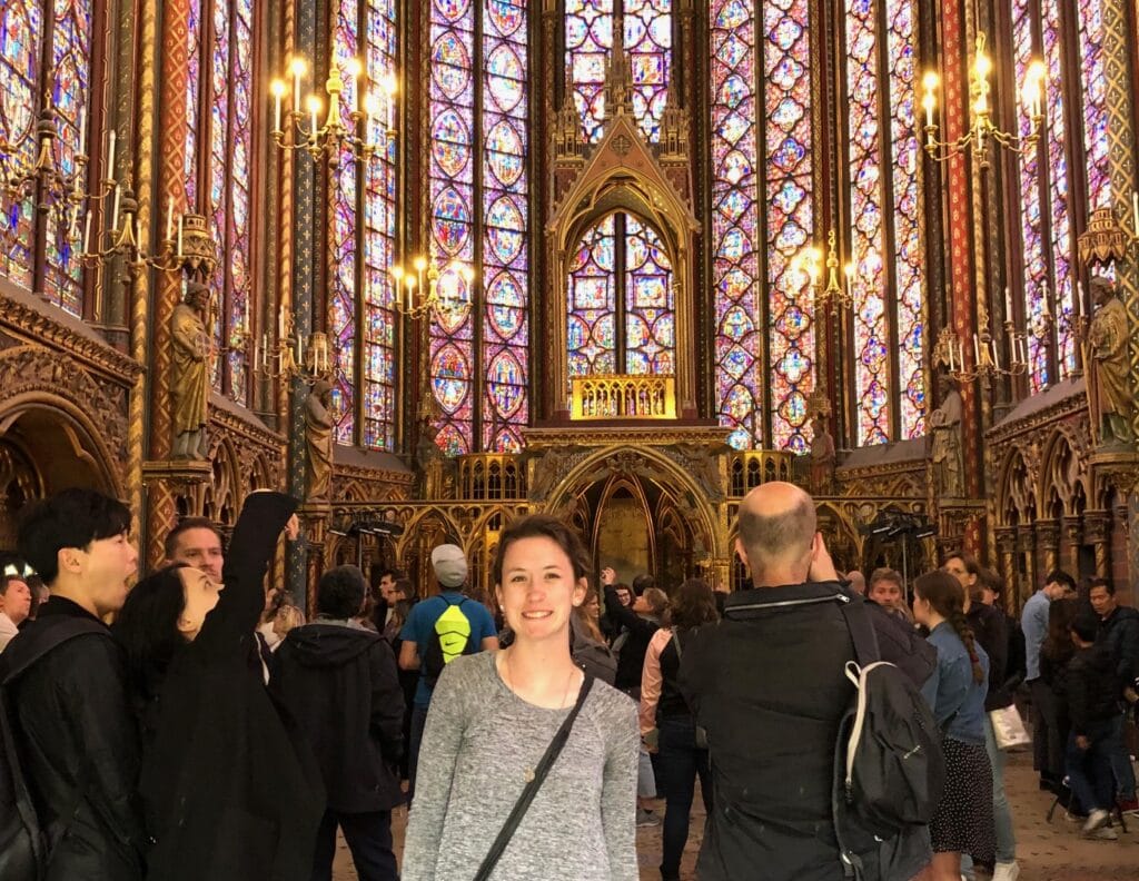 Inside the stunning Sainte-Chapelle in Paris, a woman smiles in front of vibrant, towering stained-glass windows. The ornate gothic architecture is highlighted by warm candlelight and a crowd of visitors.