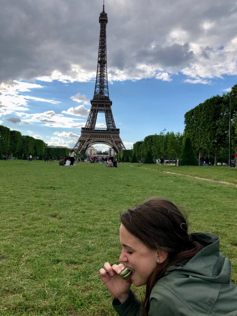  A woman enjoys a green macaron while lounging on the grass with the Eiffel Tower towering in the background under a partly cloudy sky. The scene is set in a park, with a mix of people sitting and strolling. It's the month of May which is the best month to visit Paris.