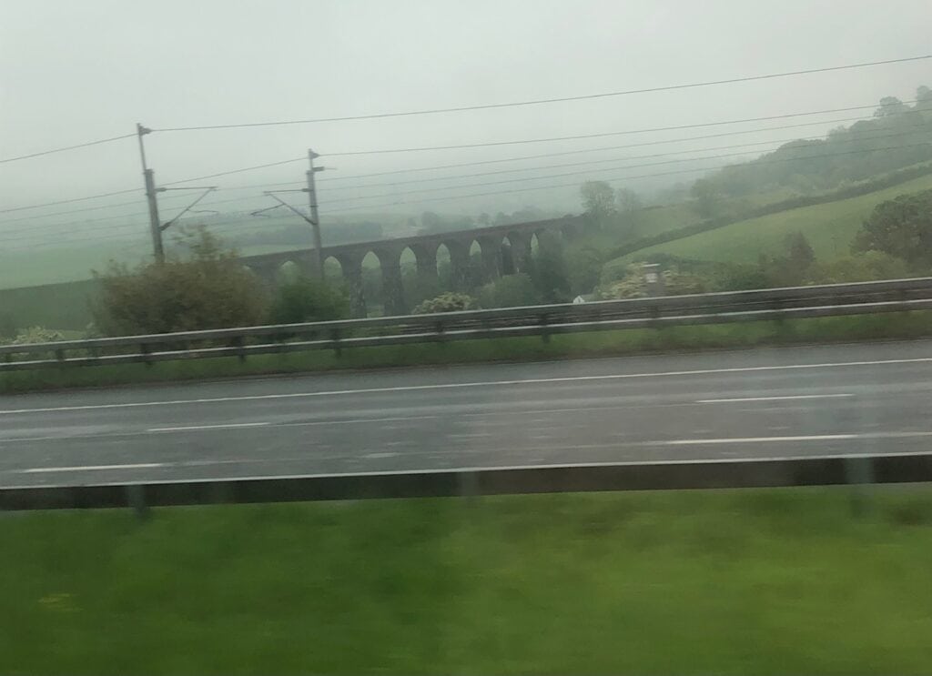 A misty, overcast landscape shows a rural scene with a multi-arched stone viaduct in the distance. The viaduct cuts across green rolling hills, while the foreground features a highway lined with a guardrail and power lines.