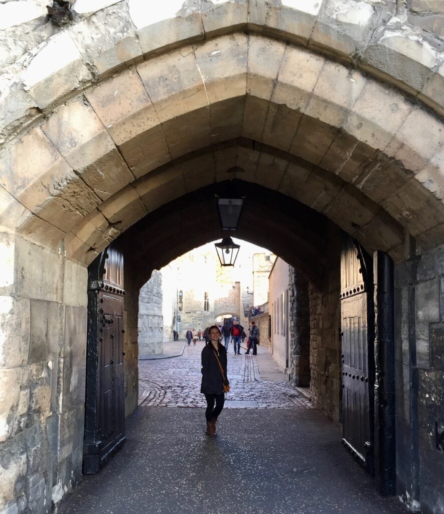 A person stands beneath a stone archway at the Tower of London. The sunlight filters through, illuminating the cobblestone street behind them, with a few other visitors visible in the distance.