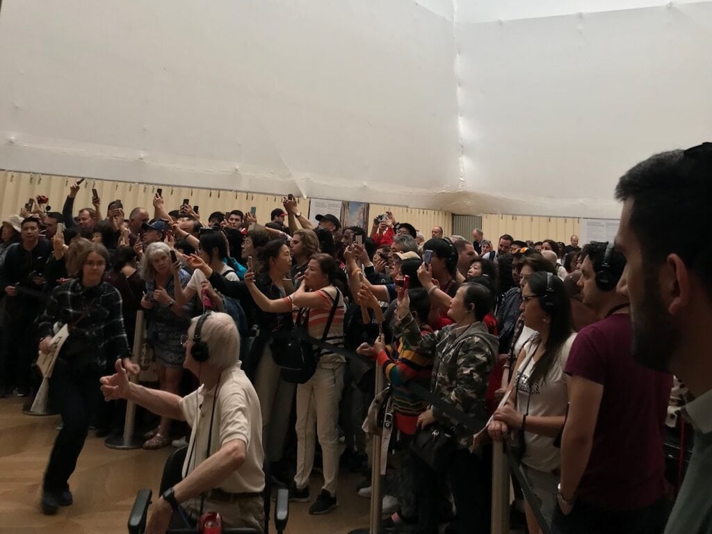 A large crowd gathers inside a museum, all focused on something outside the frame. Many people hold their phones up, likely taking pictures, and some are wearing headphones, suggesting an audio tour.