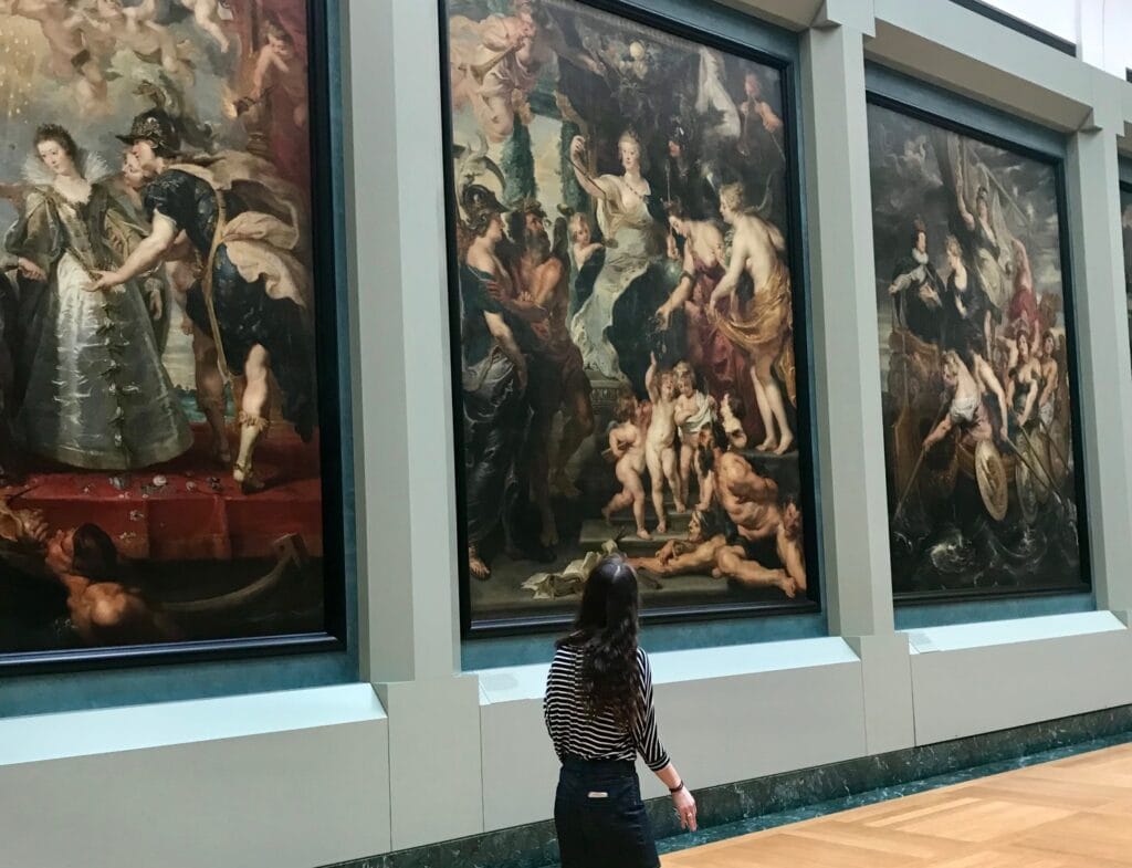 A woman stands in front of large classical paintings at the Louvre Museum, looking up at intricate, dramatic scenes depicting historical and mythological figures in vivid detail.