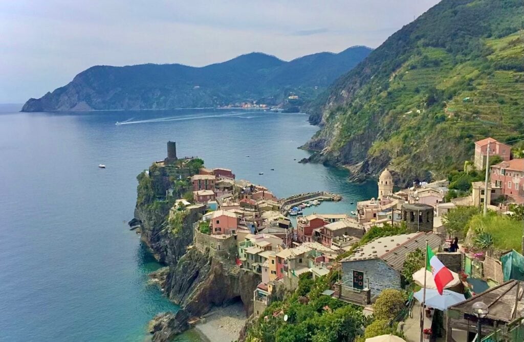 A scenic view of a coastal village perched on a cliffside. Colorful buildings are tightly packed along the rocky shore, with a small harbor visible below. Mountains rise in the background, and the ocean stretches out with calm waters. An Italian flag is prominently visible in the lower right corner, adding a national touch to the beautiful landscape.