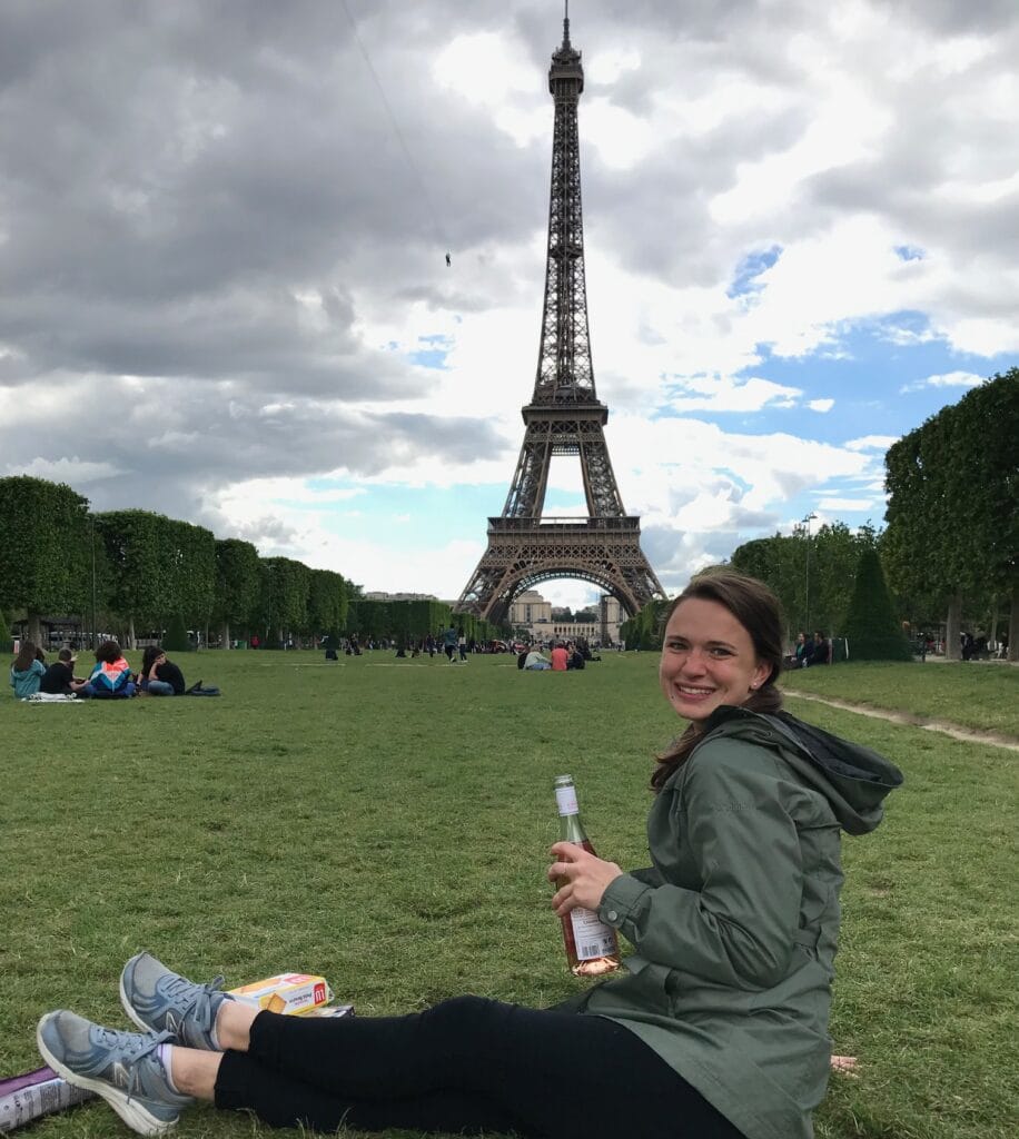 A woman sits on the grass in front of the Eiffel Tower during 2 days in Paris, smiling at the camera while holding a bottle of wine. The sky is partly cloudy, and the large open lawn is surrounded by people enjoying the view and picnicking.