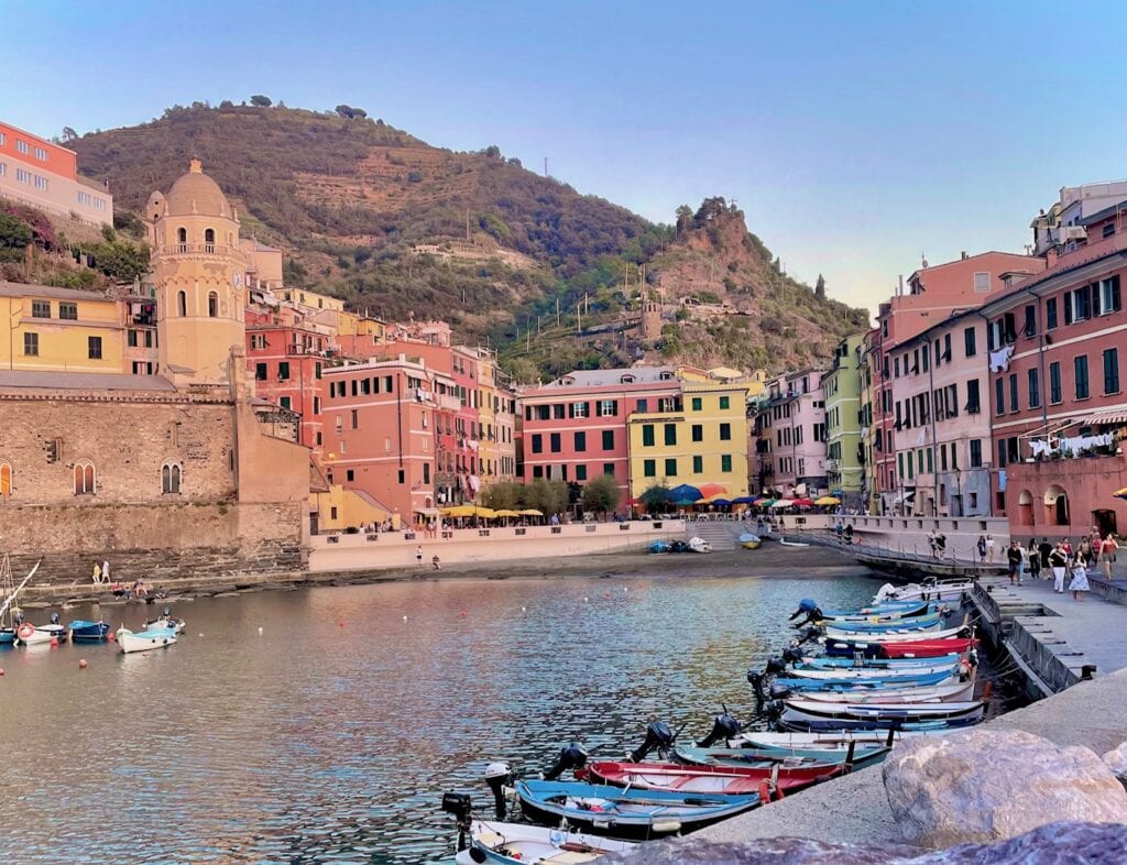 A peaceful coastal village with pastel-colored buildings nestled along a small harbor. The water is calm, and several small boats are docked near the shoreline. In the background, a hillside covered with greenery rises behind the village, giving a picturesque view of the town at the foot of the hill.