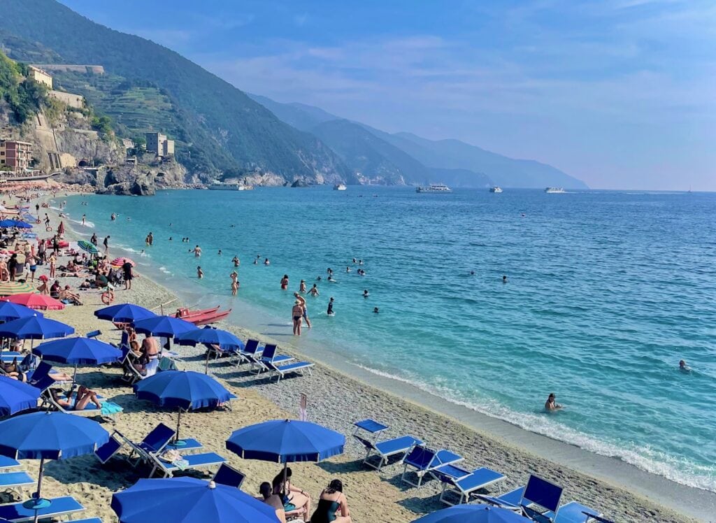 A picturesque beach scene with bright blue water and many beachgoers enjoying the sea. Blue beach umbrellas and lounge chairs are scattered across the sandy shore. In the background, green hills slope down towards the coast, and boats can be seen floating on the water. The sky is clear and sunny, with distant hills fading into the horizon.