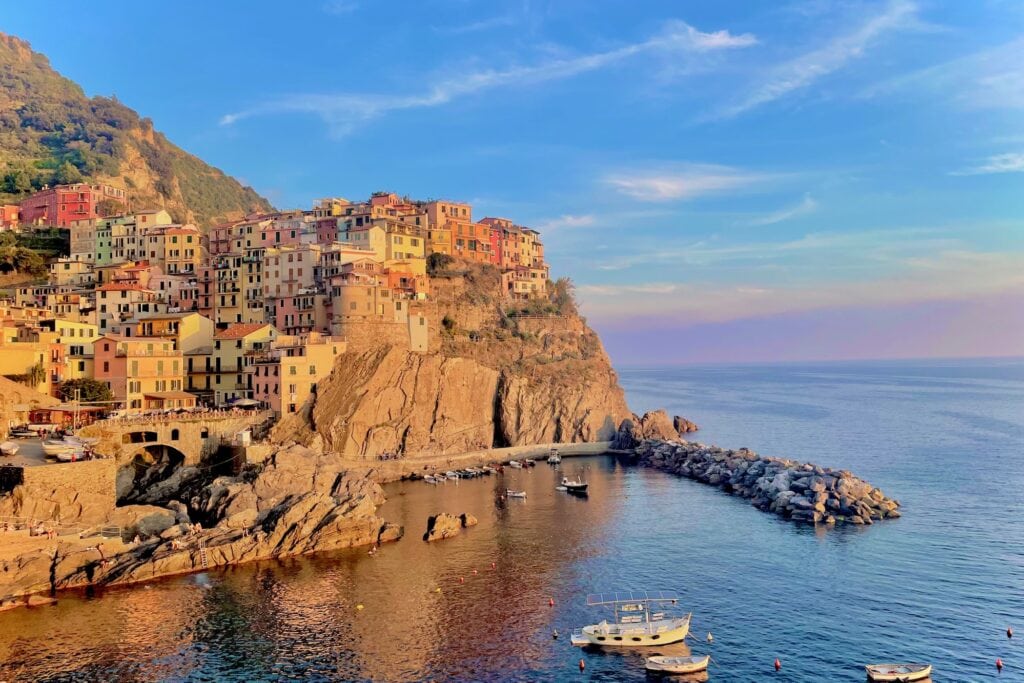 A stunning view of a cliffside town with colorful buildings stacked on top of one another, overlooking the calm, clear blue sea. A few small boats are anchored near the rocky coastline, with the golden hues of sunset casting a warm glow on the entire scene.