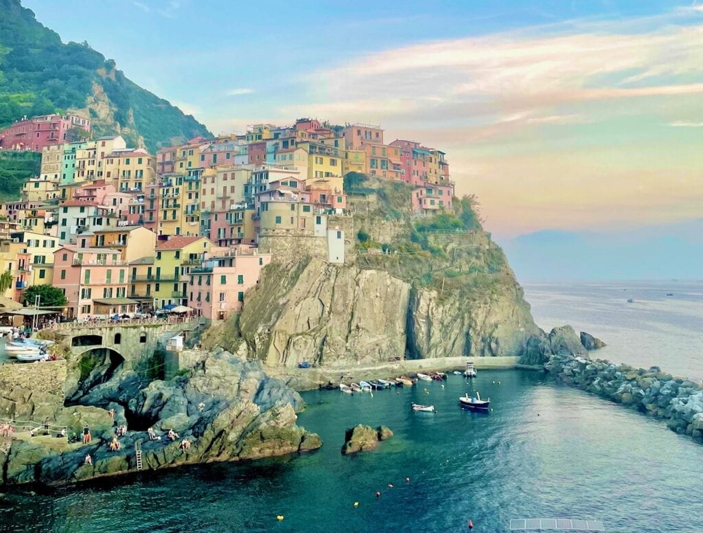 A sunset view of a colorful Italian cliffside village with vibrant houses stacked on steep rocky slopes. Below, the calm sea reflects the warm hues of the sky, and small boats float near the harbor.