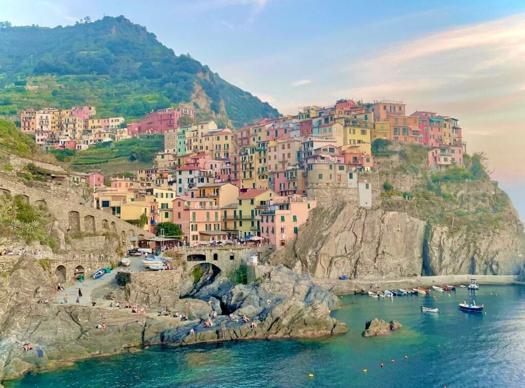 A cliffside village scene with multicolored houses perched above the sea. The rocky coastline features swimmers enjoying the water, and small boats are moored in a small, protected bay. The scene is captured during the golden hour, highlighting the warm tones of the buildings and landscape.