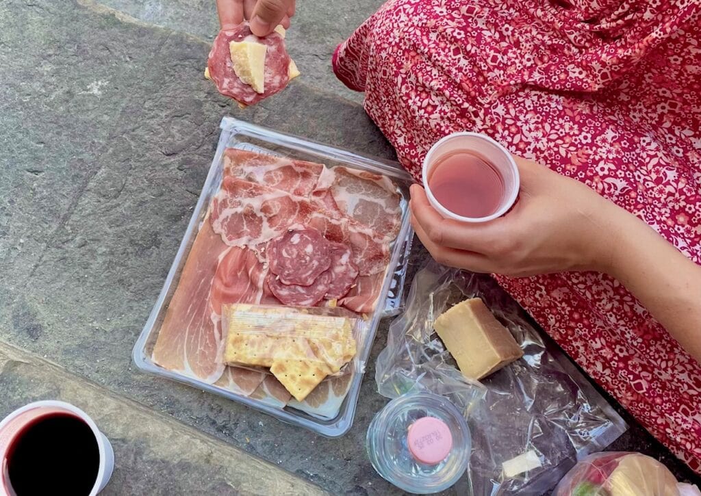 A person in a red floral dress is having a picnic. There's a plastic tray filled with various cured meats like prosciutto, salami, and slices of cheese. Crackers are stacked on top of the meats, and a hand is seen holding a piece of salami with a chunk of cheese on top. Another hand holds a cup filled with a pink drink, possibly wine. There's also a plastic container with a clear bottle cap visible in the lower corner.