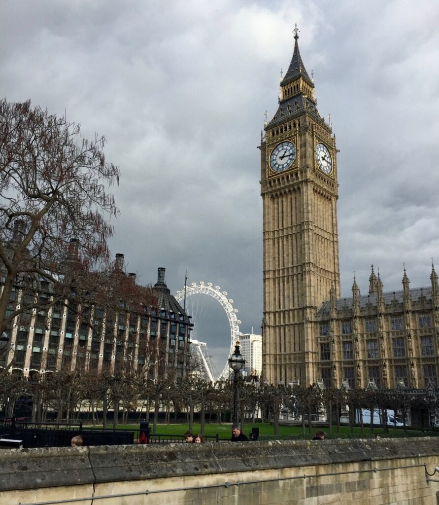 Big Ben stands tall in the foreground with the London Eye visible in the background. The sky is cloudy, and bare trees line the scene, with buildings and part of the Palace of Westminster in view.