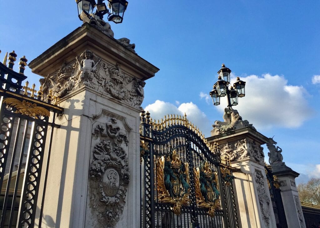 Close-up of the intricately decorated gates of Buckingham Palace, featuring ornate stone carvings, golden accents, and regal emblems, all under a bright blue sky.