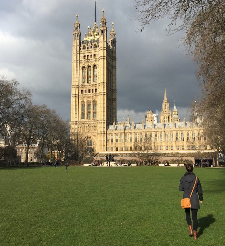 A woman following this packing list for London in winter wearing a gray coat and leather purse walks along a grassy area in front of London's Parliament building.